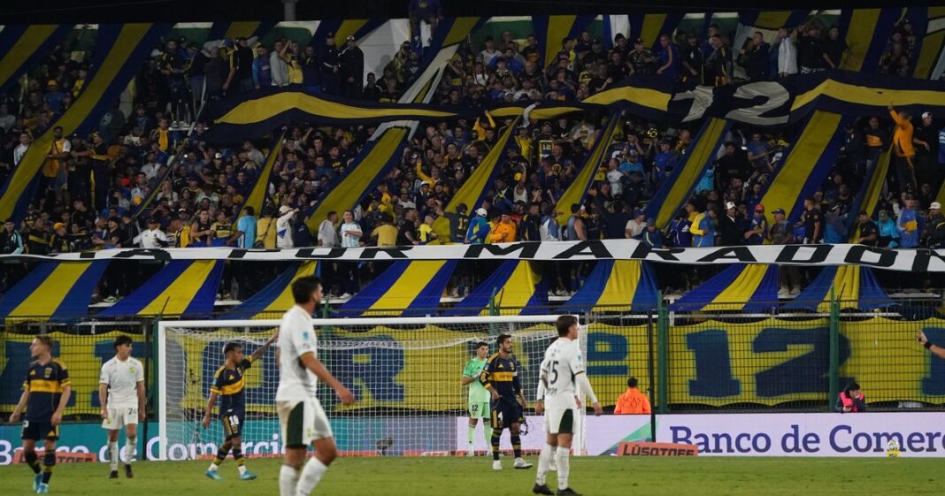 Hinchas visitantes en un estadio de fútbol argentino, con banderas y camisetas, durante un partido de la Liga Profesional