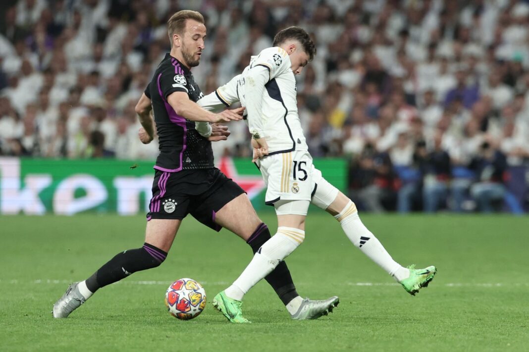 Los escudos del Real Madrid y el Bayern Munich enfrentados, con el trofeo de la Champions League de fondo.