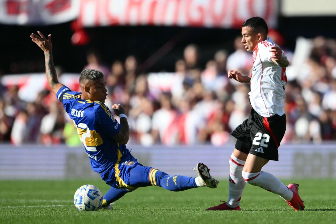 Escudos de River Plate y Boca Juniors enfrentados, con el estadio Monumental de fondo.